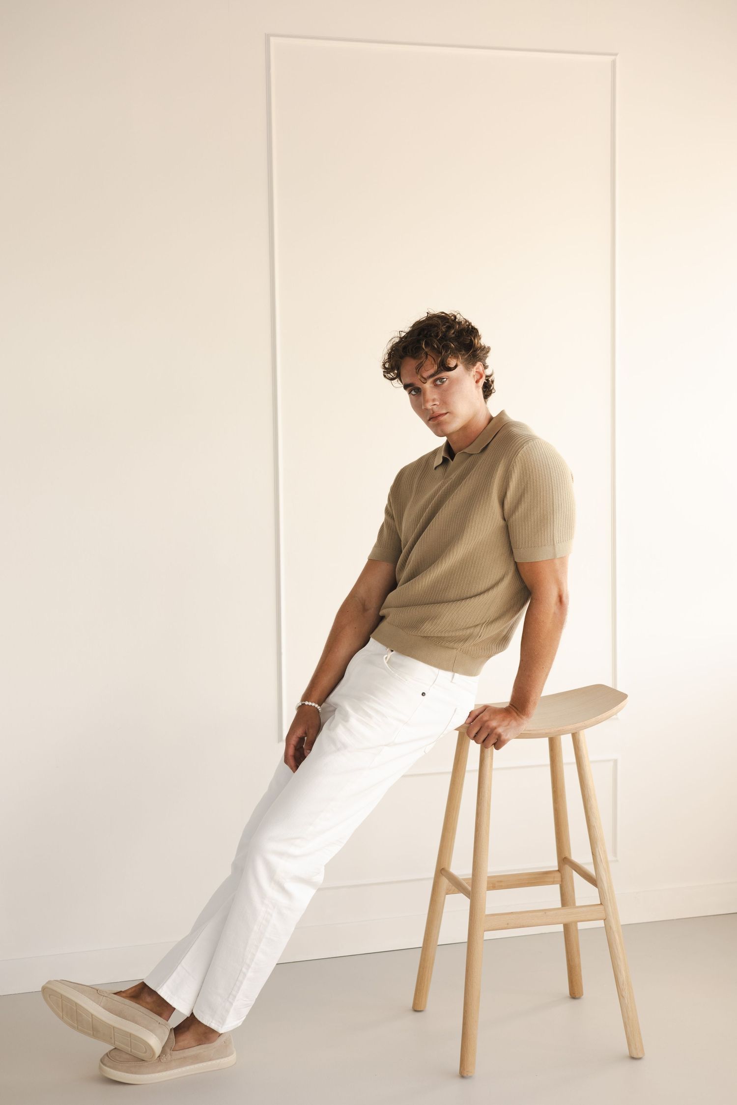 Young man in beige polo shirt and white pants sitting on a wooden bar stool.