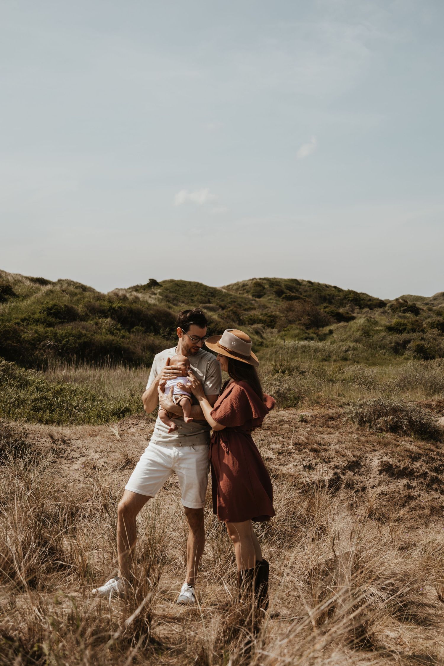 A couple stands in a golden grass field holding their newborn baby under a bright sky.