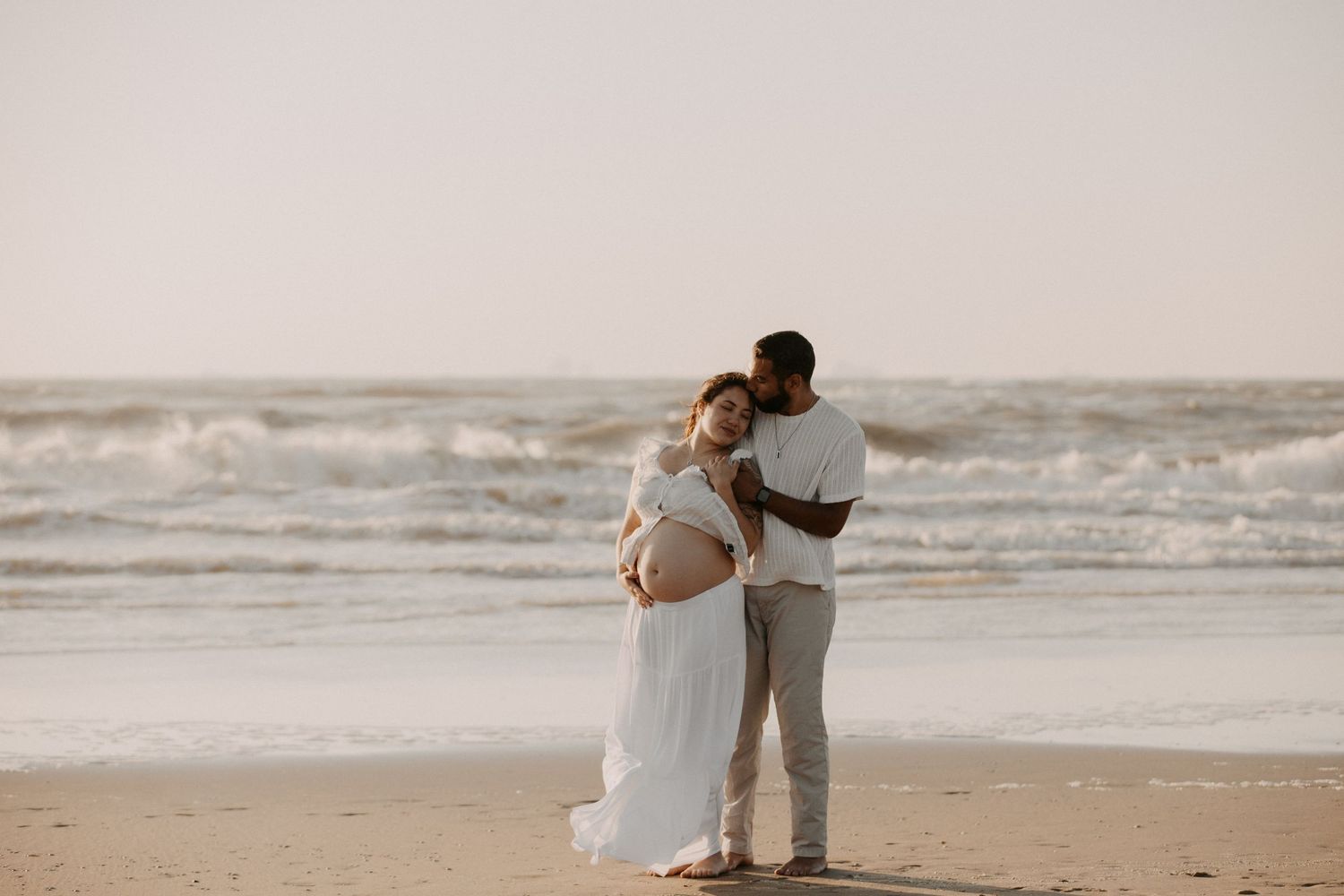 Pregnant couple embracing on a beach at sunset, woman in white dress cradling baby bump with ocean waves behind them.