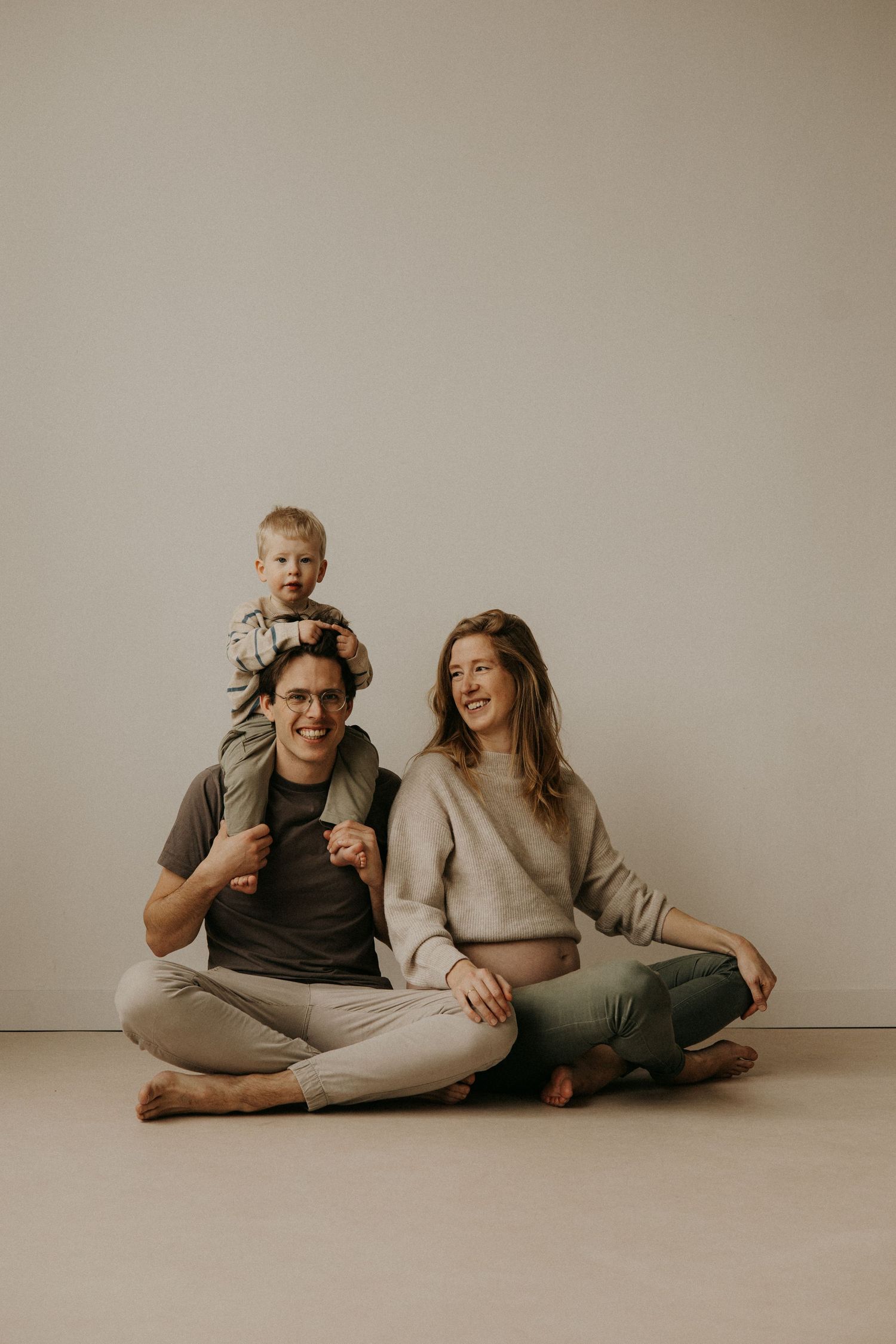 A young family with a toddler on dad's shoulders sitting cross-legged on the floor during a maternity photo session.
