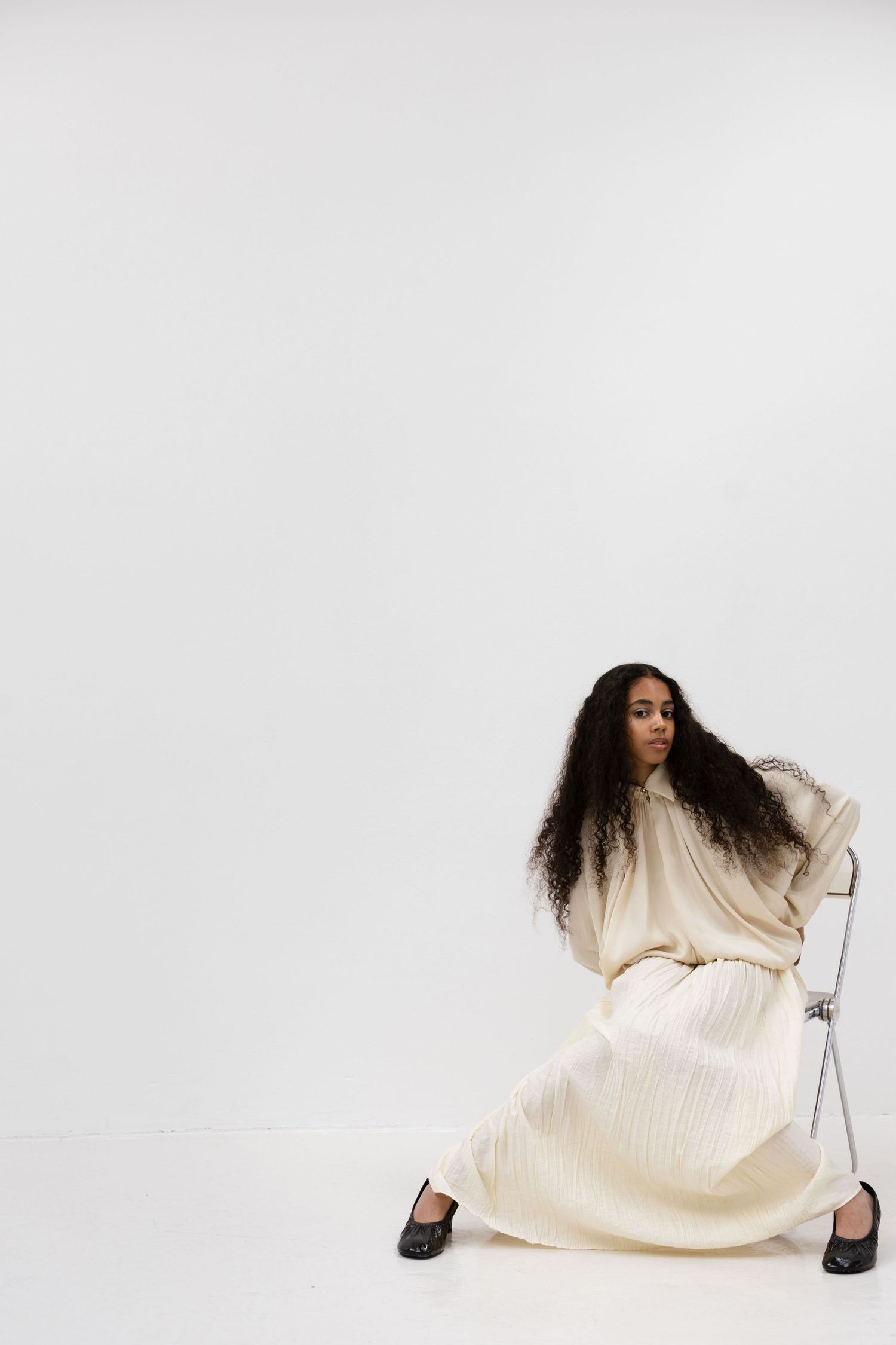 Woman in cream pleated maxi dress and black loafers posing on chair against white studio background.