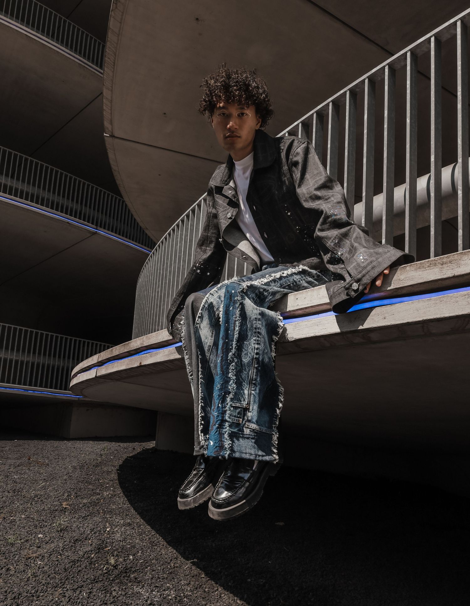 Man with curly hair sitting on a concrete ledge wearing a dark coat and wide-leg jeans.