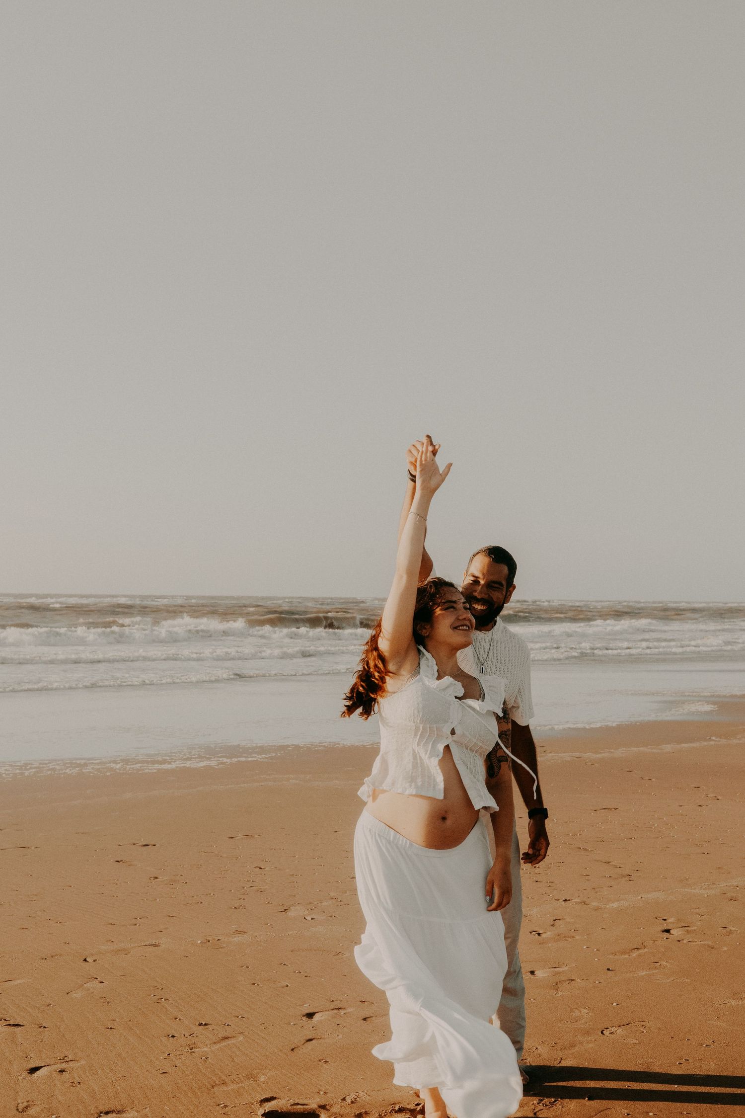 Pregnant couple dancing joyfully on a sandy beach, woman in white dress with arm raised, man in white shirt behind her.