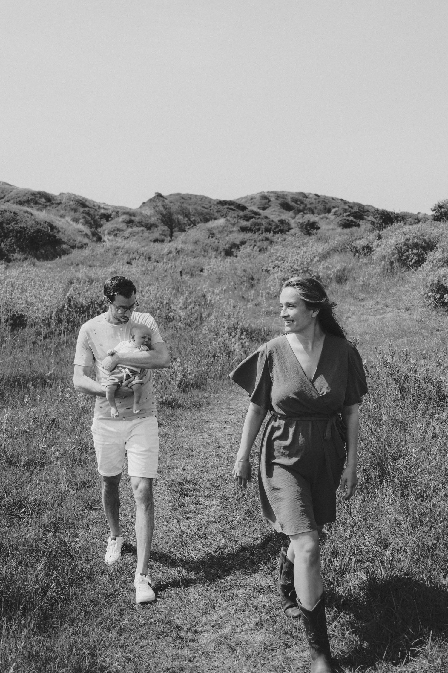 Black and white photo of a couple walking a grassy path, man carrying a newborn baby.