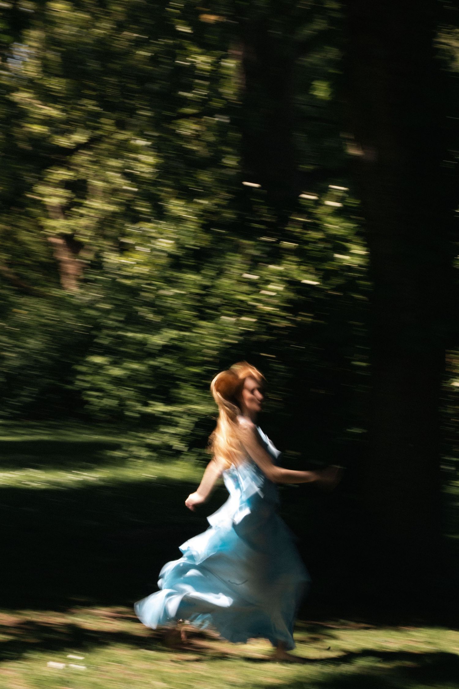 Motion blur of woman in light blue dress running through sunlit green forest.