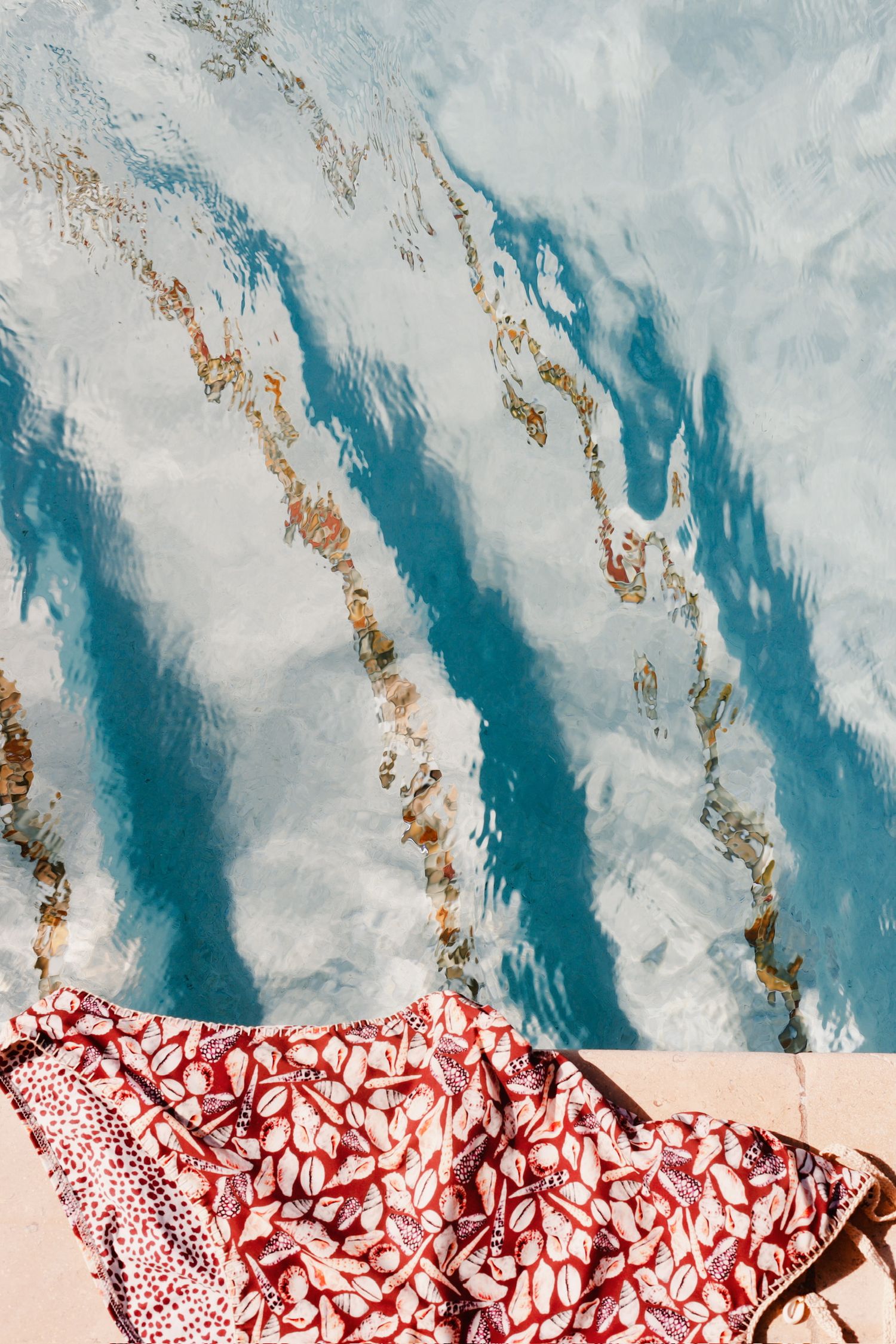 Aerial view of a red floral print swimsuit coverup laid on pool edge with blue rippling water.