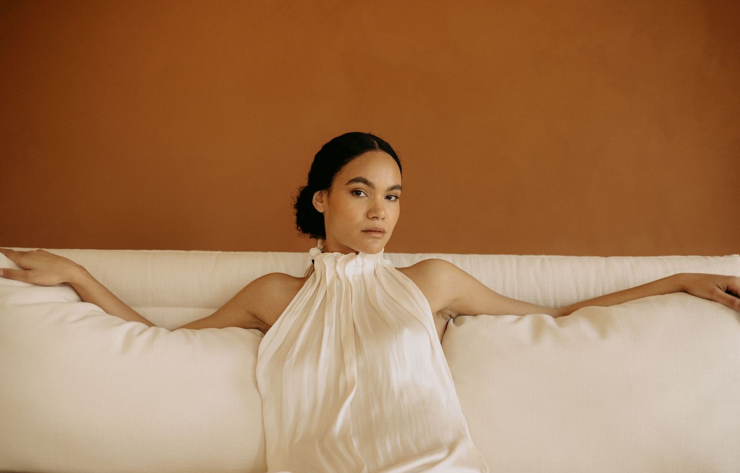 Woman in white halter dress reclines on a cream sofa against a warm terracotta wall.