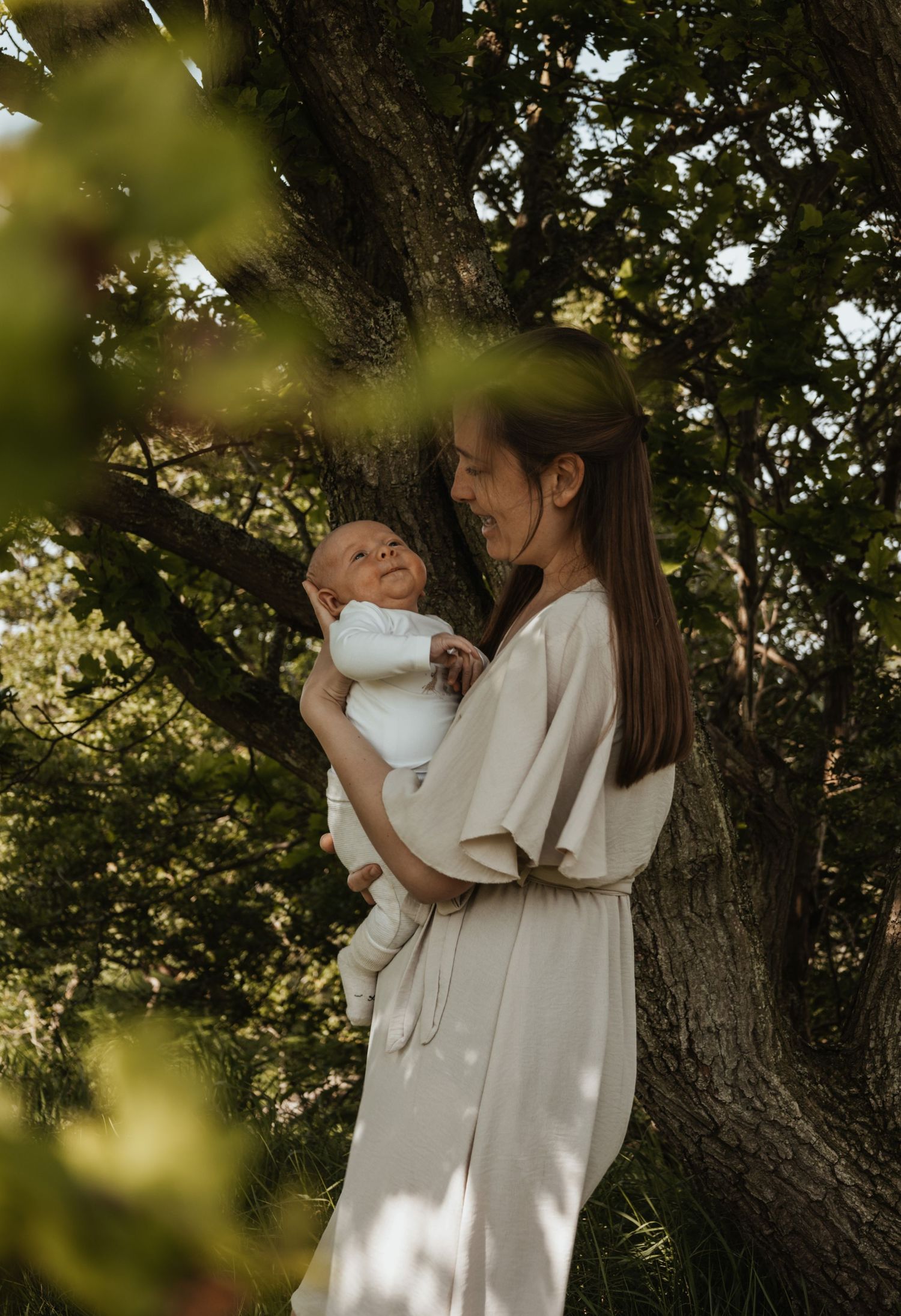 A smiling mother holds her newborn baby while leaning against a large tree in a sunny garden.
