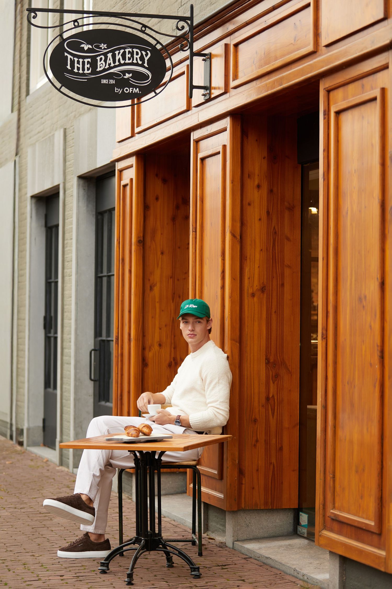 Man in white outfit and green cap sitting outside The Bakery cafe with croissants.