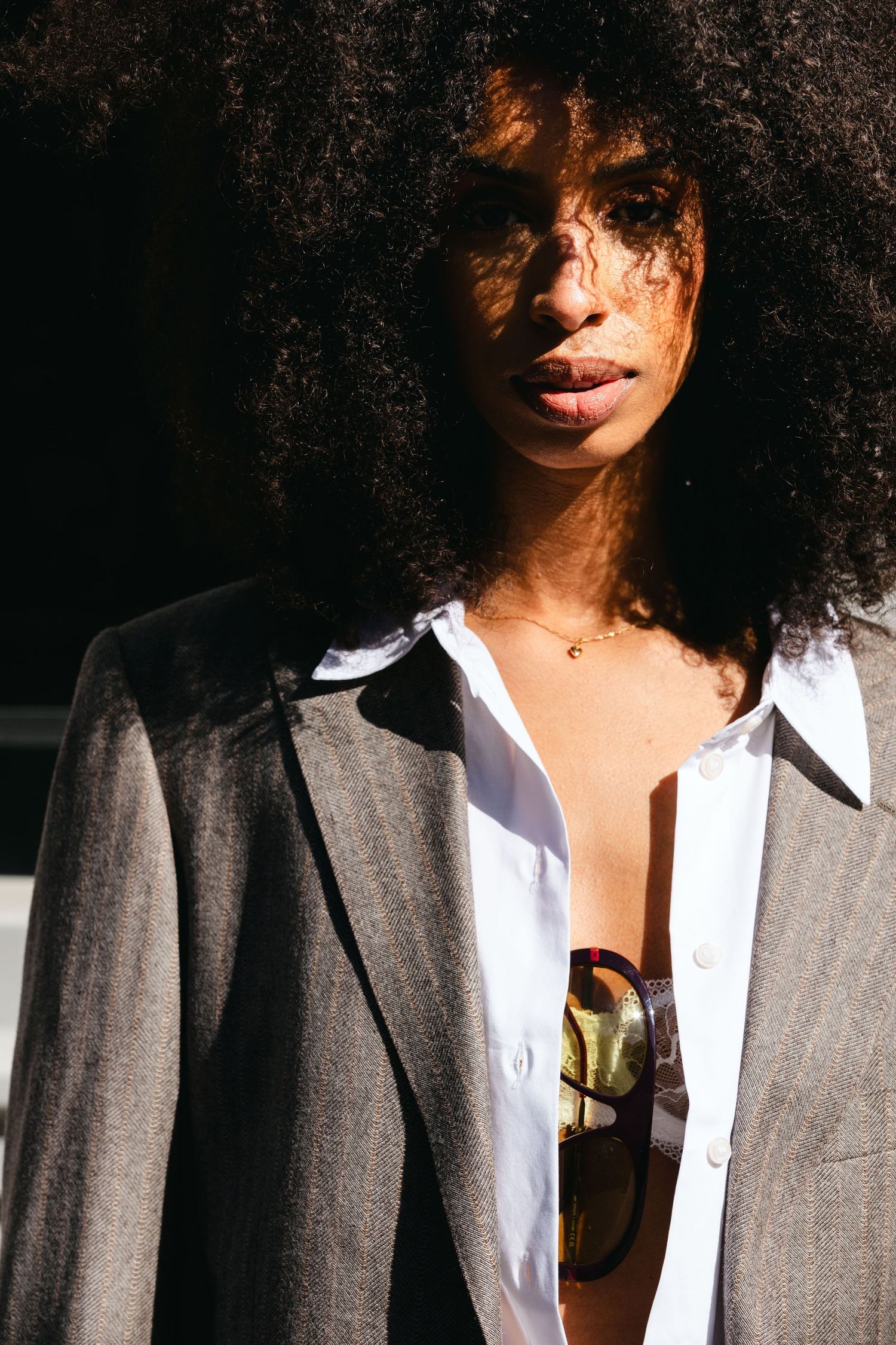 Woman with voluminous afro hair in grey blazer and open white shirt poses in bright sunlight.