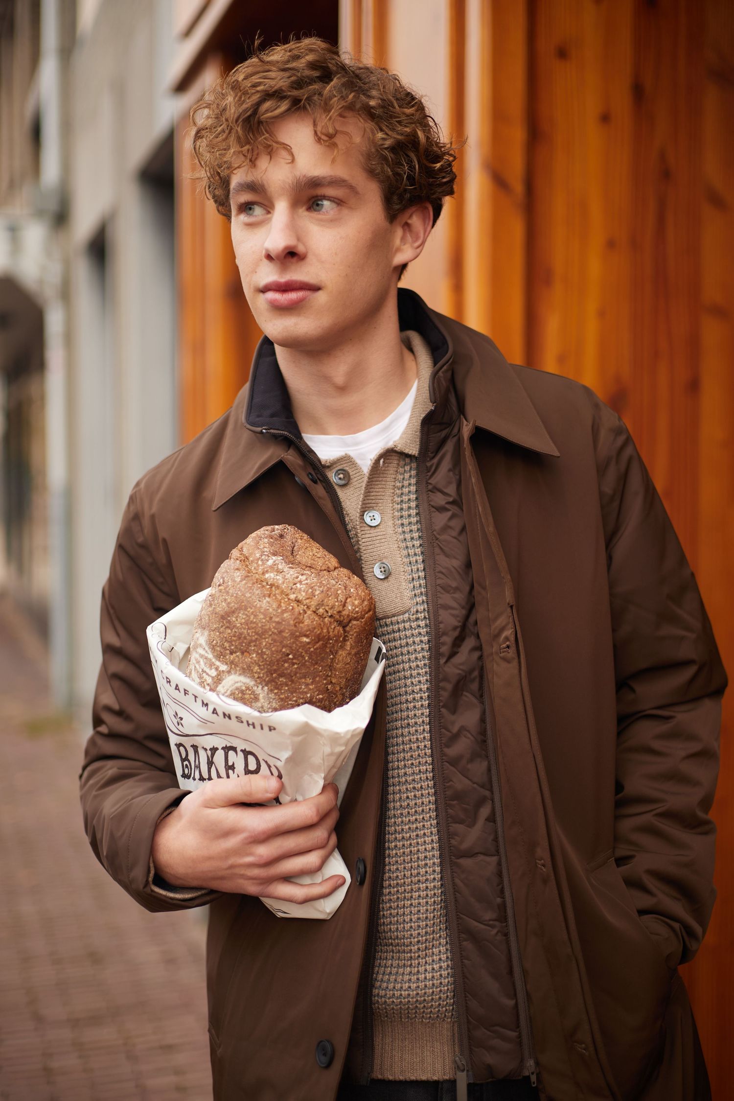 Young man in brown quilted jacket holding artisan bakery bread loaf on city street.