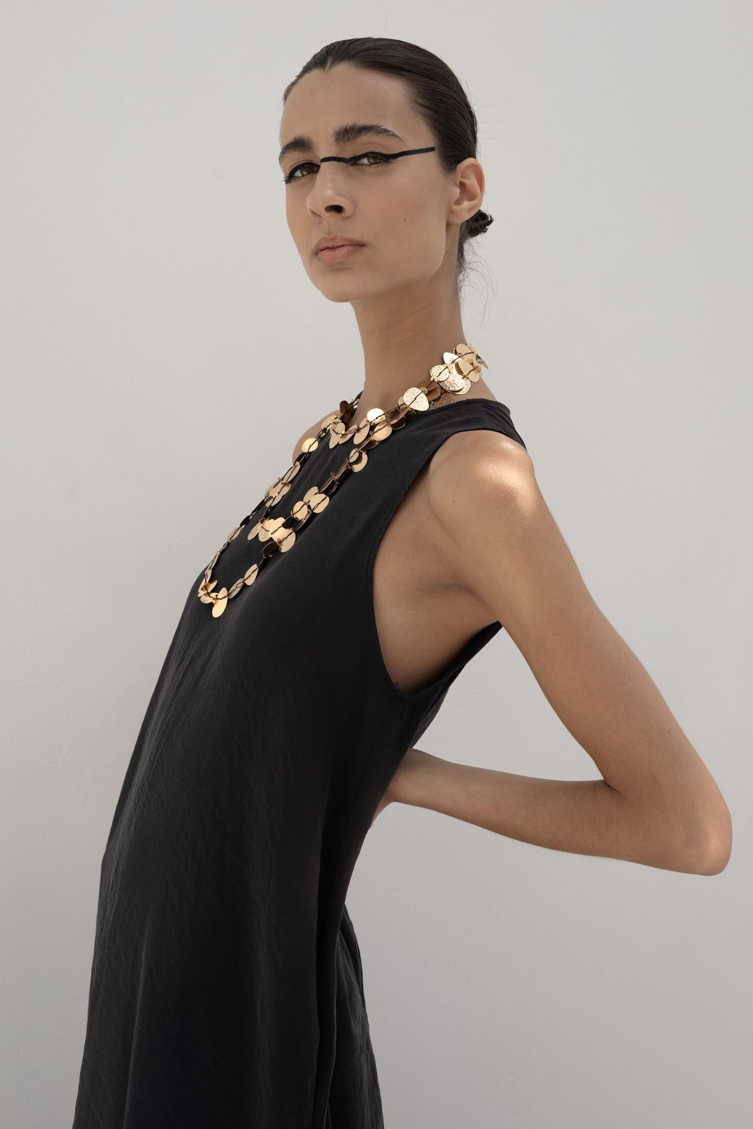 Model in black sleeveless dress wearing gold coin chain necklace posing in studio with sleek hair.