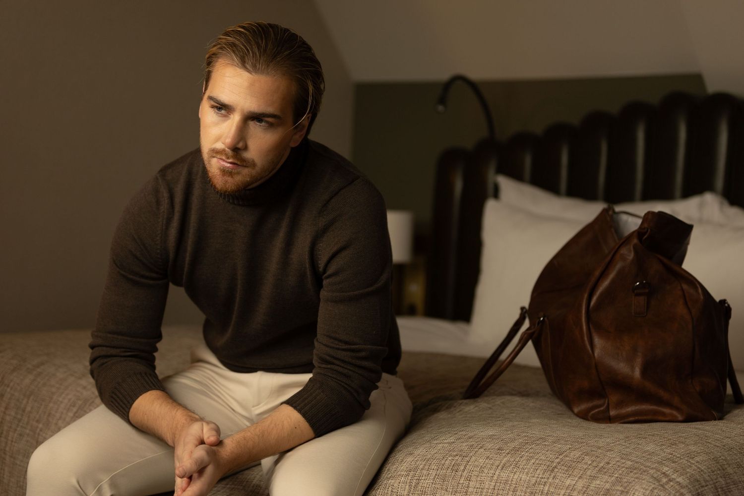 Man in brown sweater sits on hotel bed beside large brown leather duffle bag.