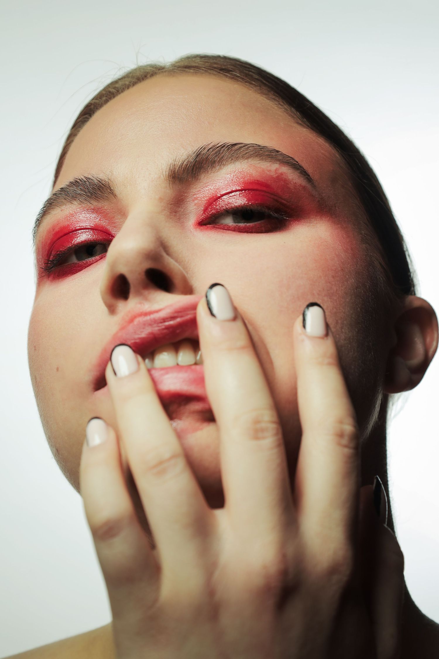 Close-up portrait of a woman with bold red eyeshadow, white nails, pressing hands against her face.