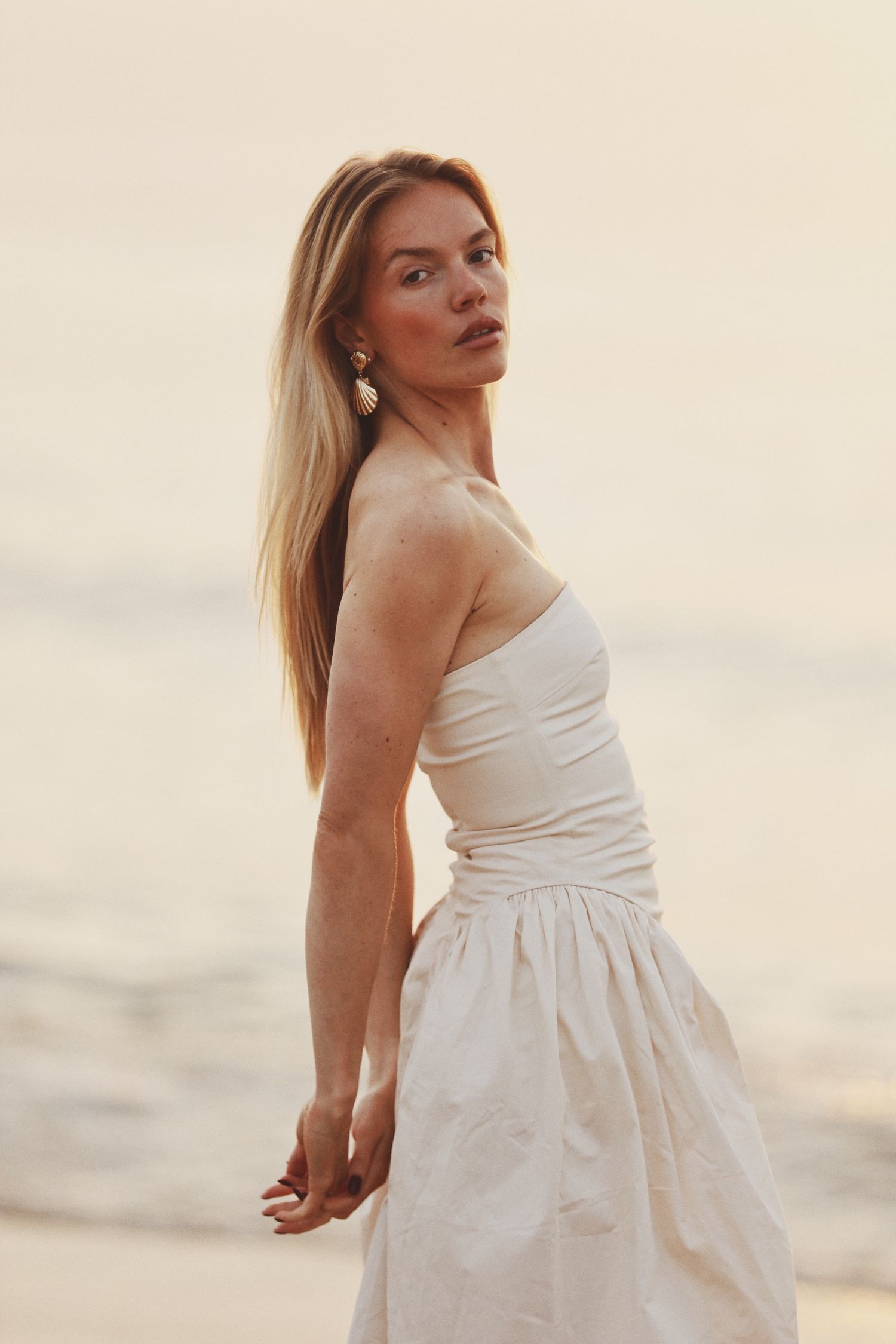 Blonde woman in a white strapless dress poses gracefully on a beach at golden hour sunset.