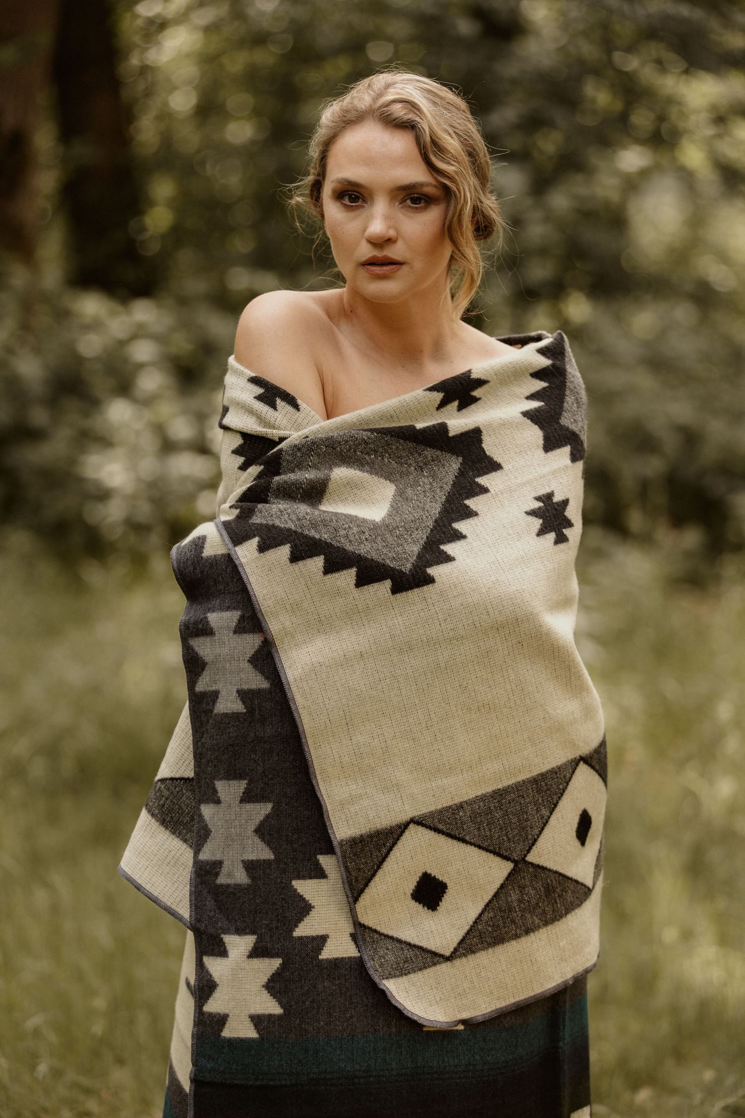Woman wrapped in a geometric patterned blanket standing outdoors in a lush green forest setting.