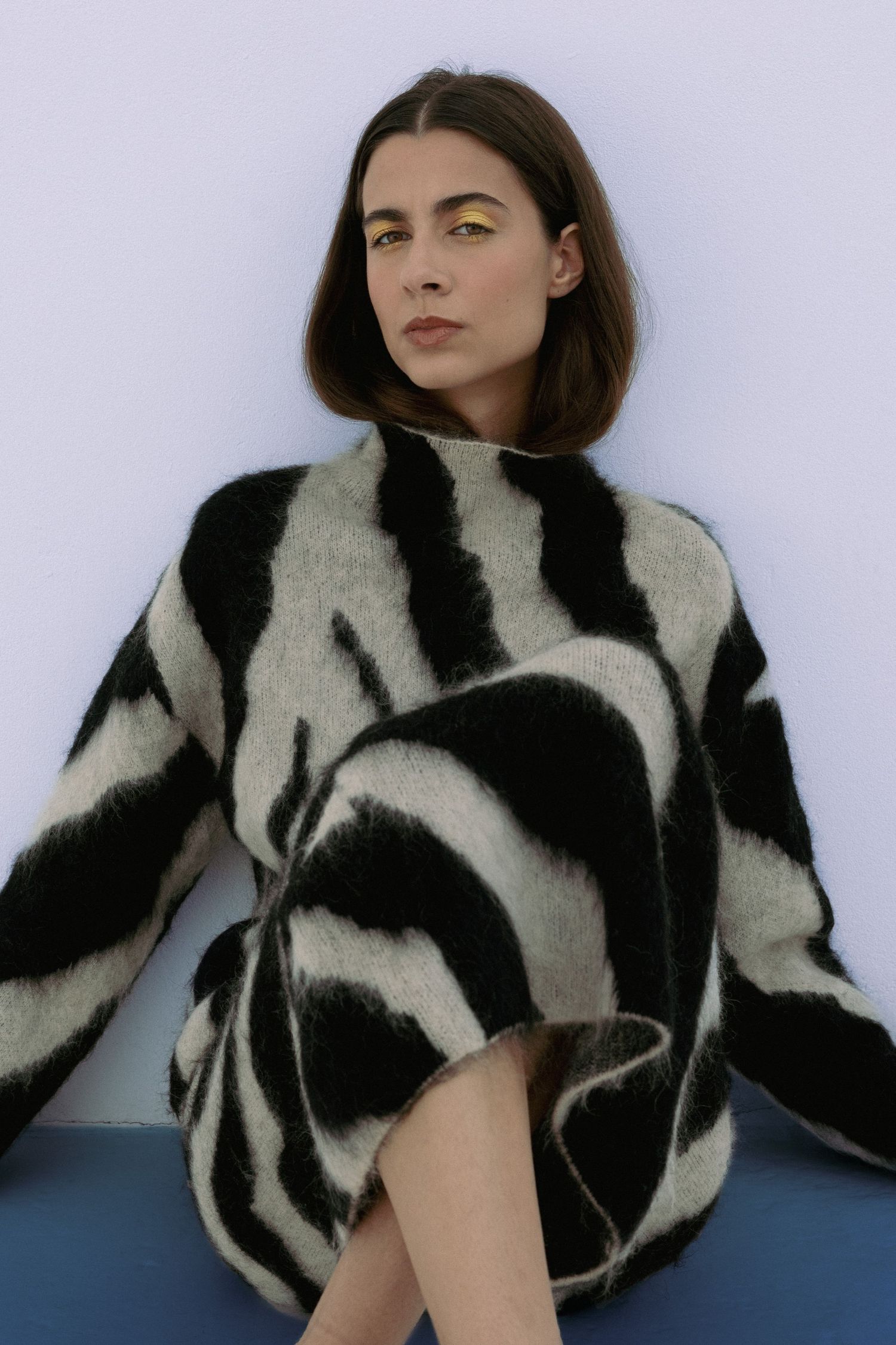 Brunette woman in oversized black and white zebra print fuzzy sweater sitting against white wall.