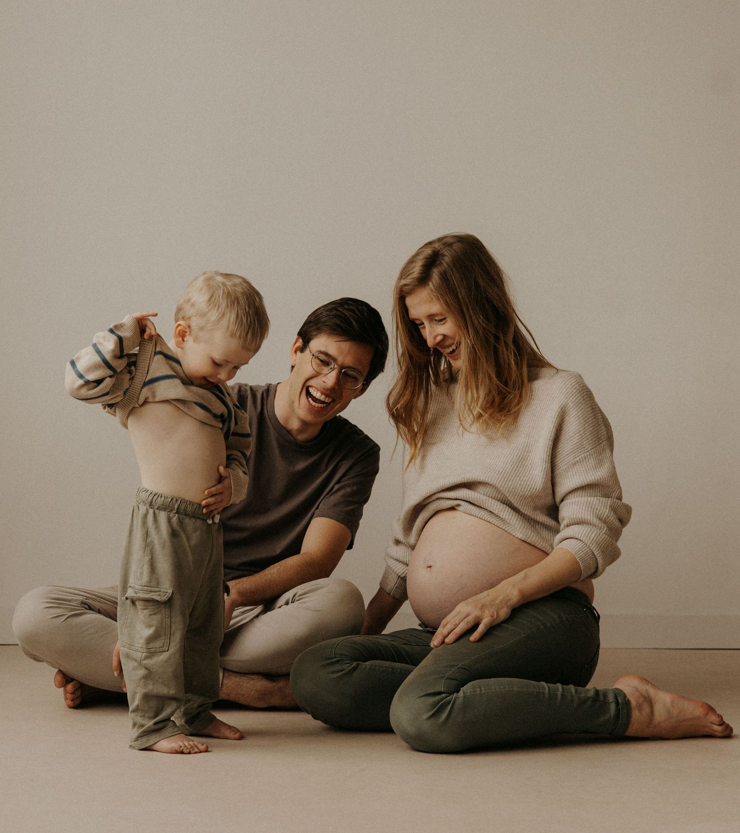 A laughing family with a playful toddler lifting his shirt during a casual maternity photoshoot on the floor.