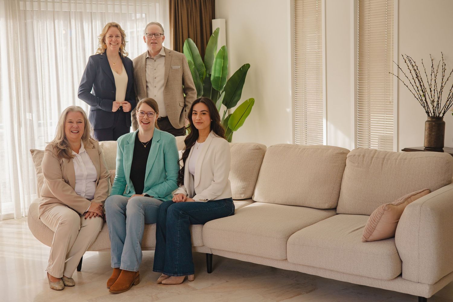Five professional colleagues posing together in a modern living room with a beige sofa and elegant decor.