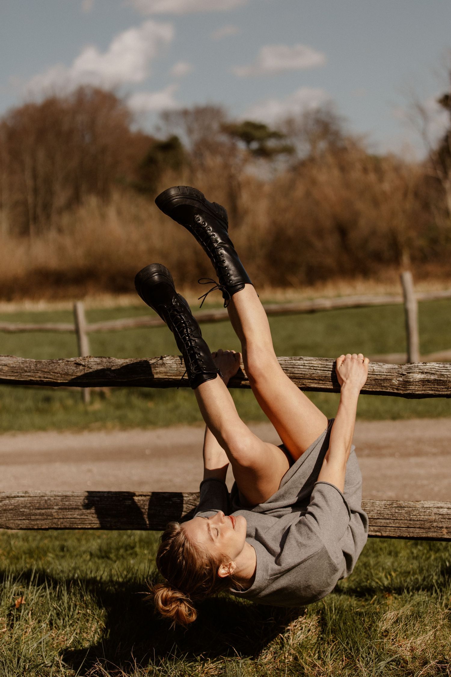 Woman in gray dress and black boots lying on grass beside rustic wooden fence outdoors.