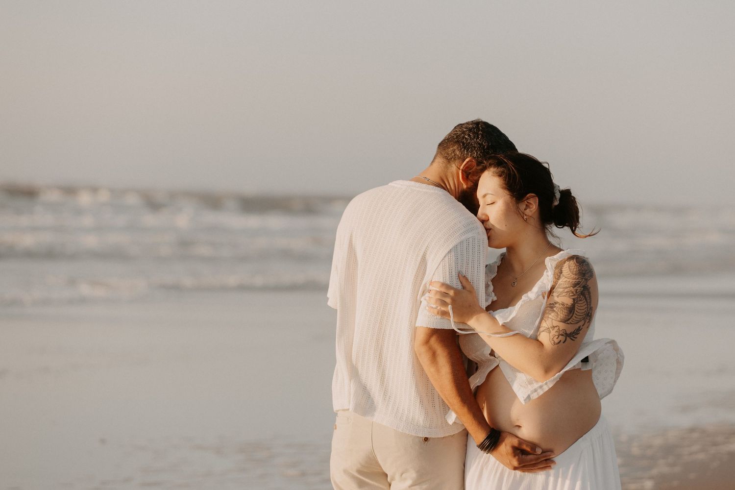 Pregnant couple embracing tenderly on a beach at sunset, woman with tattoos in white dress, waves in background.