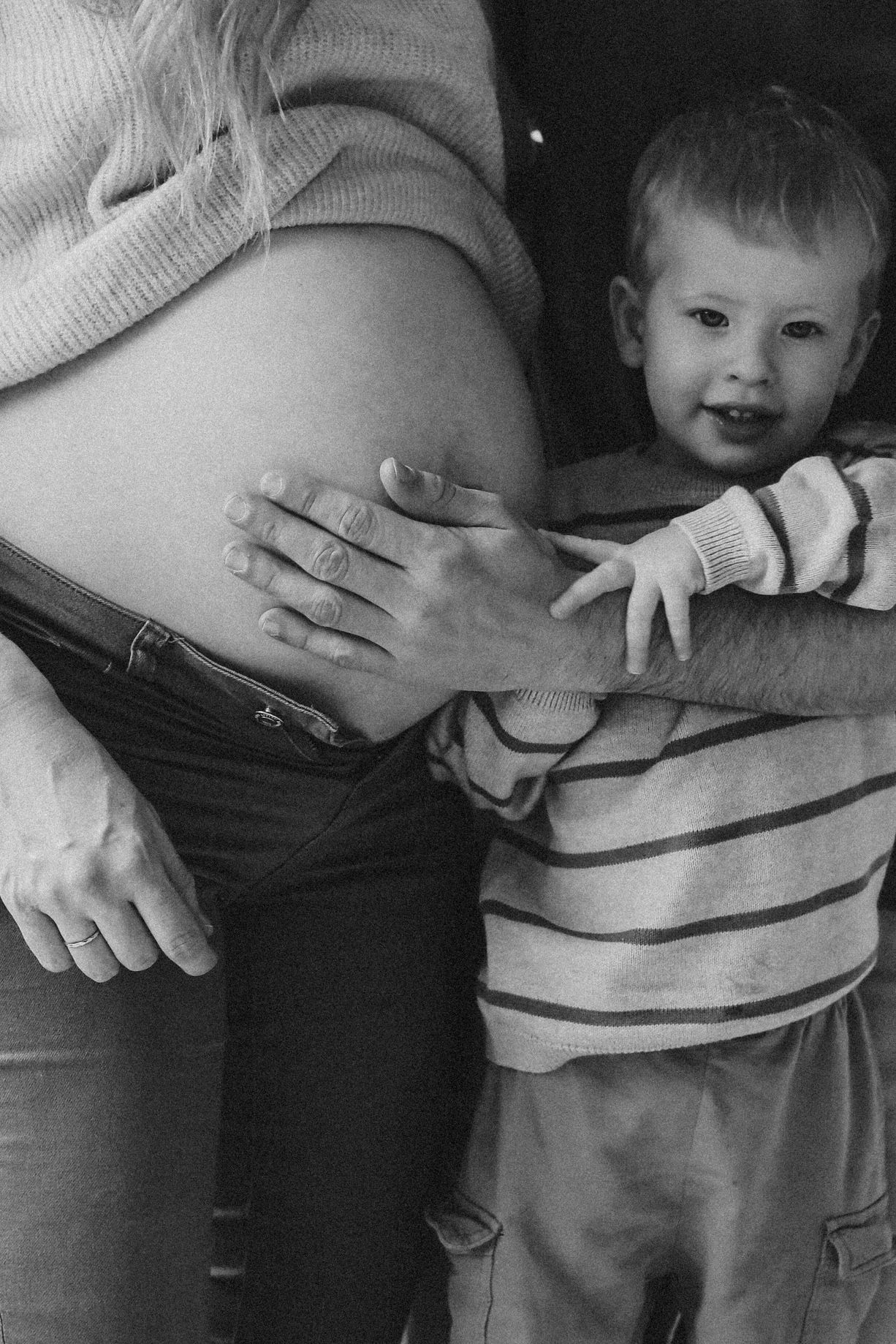 Black and white photo of a toddler hugging a pregnant mother's baby bump in an intimate maternity portrait.