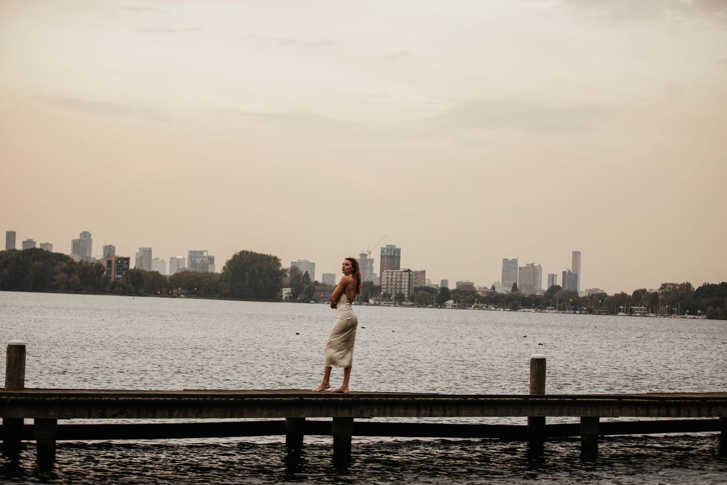 Woman in a white dress stands on a wooden pier with a city skyline and river at sunset.