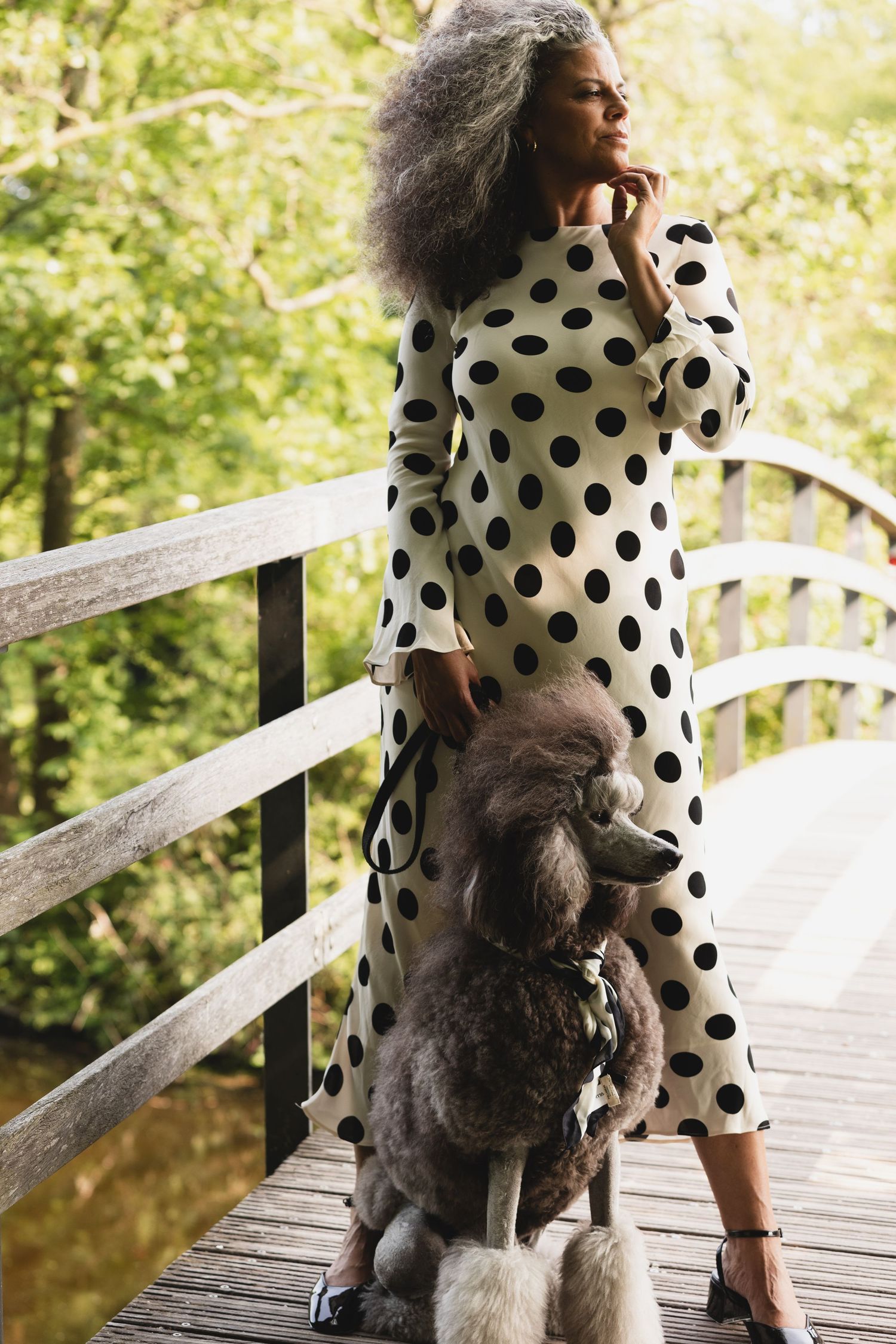 Woman in black and white polka dot dress with grey poodle on wooden bridge.