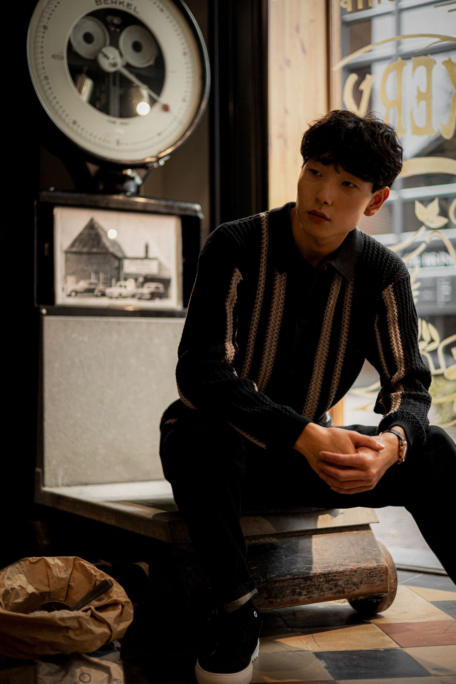 Young man in striped black sweater sits on wooden bench in vintage shop with antique scale.