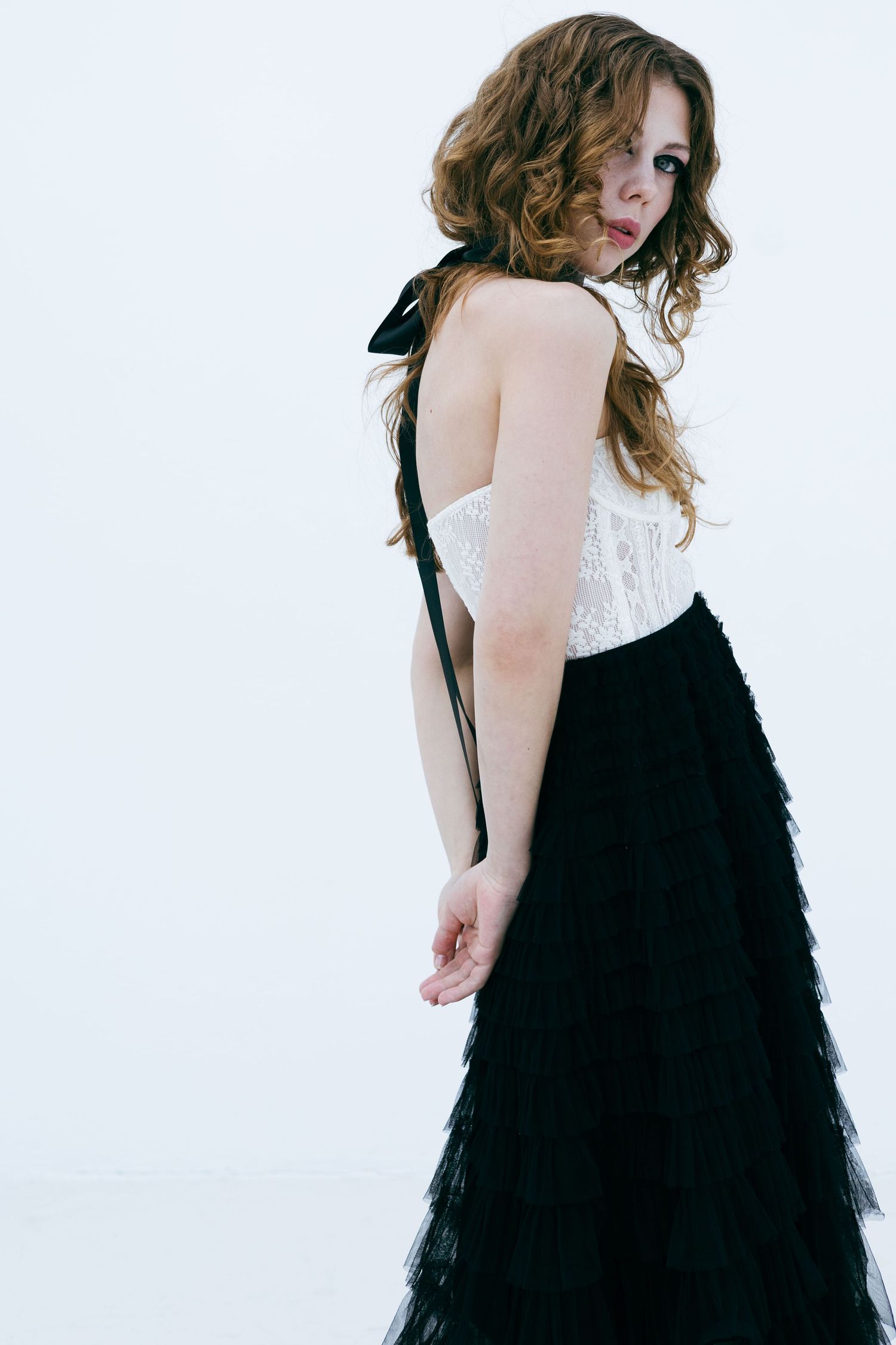 Woman with curly auburn hair in white lace top and black ruffled skirt on white background.