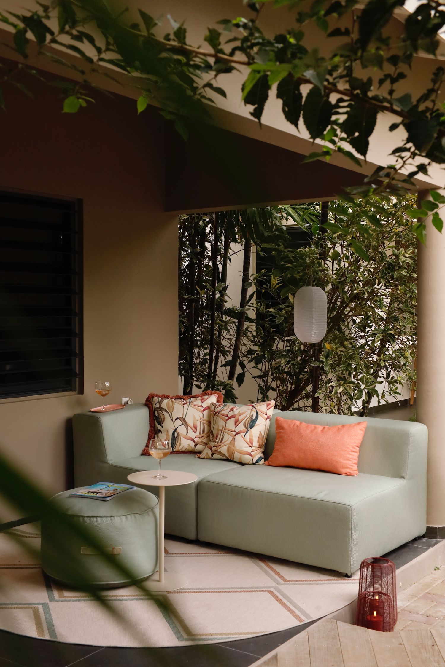 Sage green outdoor sofa with floral and coral cushions on shaded patio surrounded by plants.