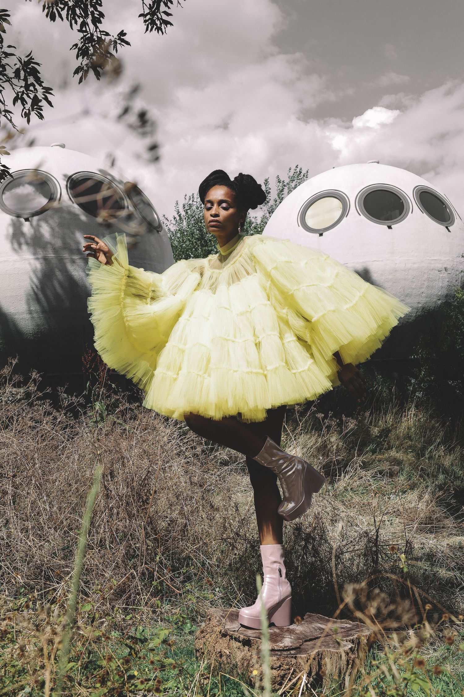 Woman in yellow tulle dress posing outdoors near futuristic dome structures wearing platform heels.