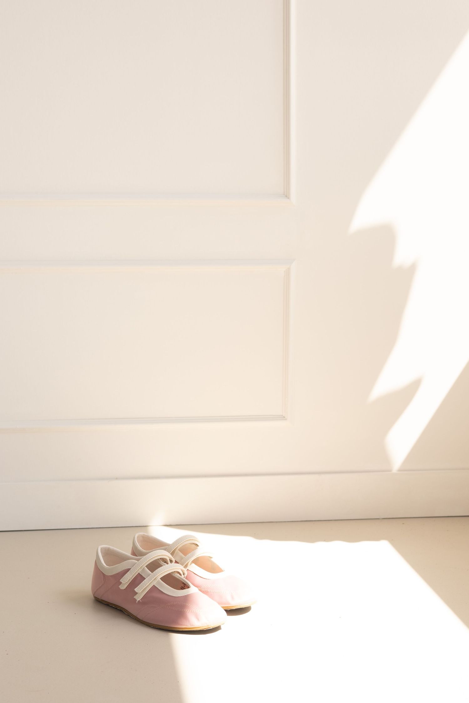 Small pink shoes with white straps on light floor against white paneled wall in soft sunlight.