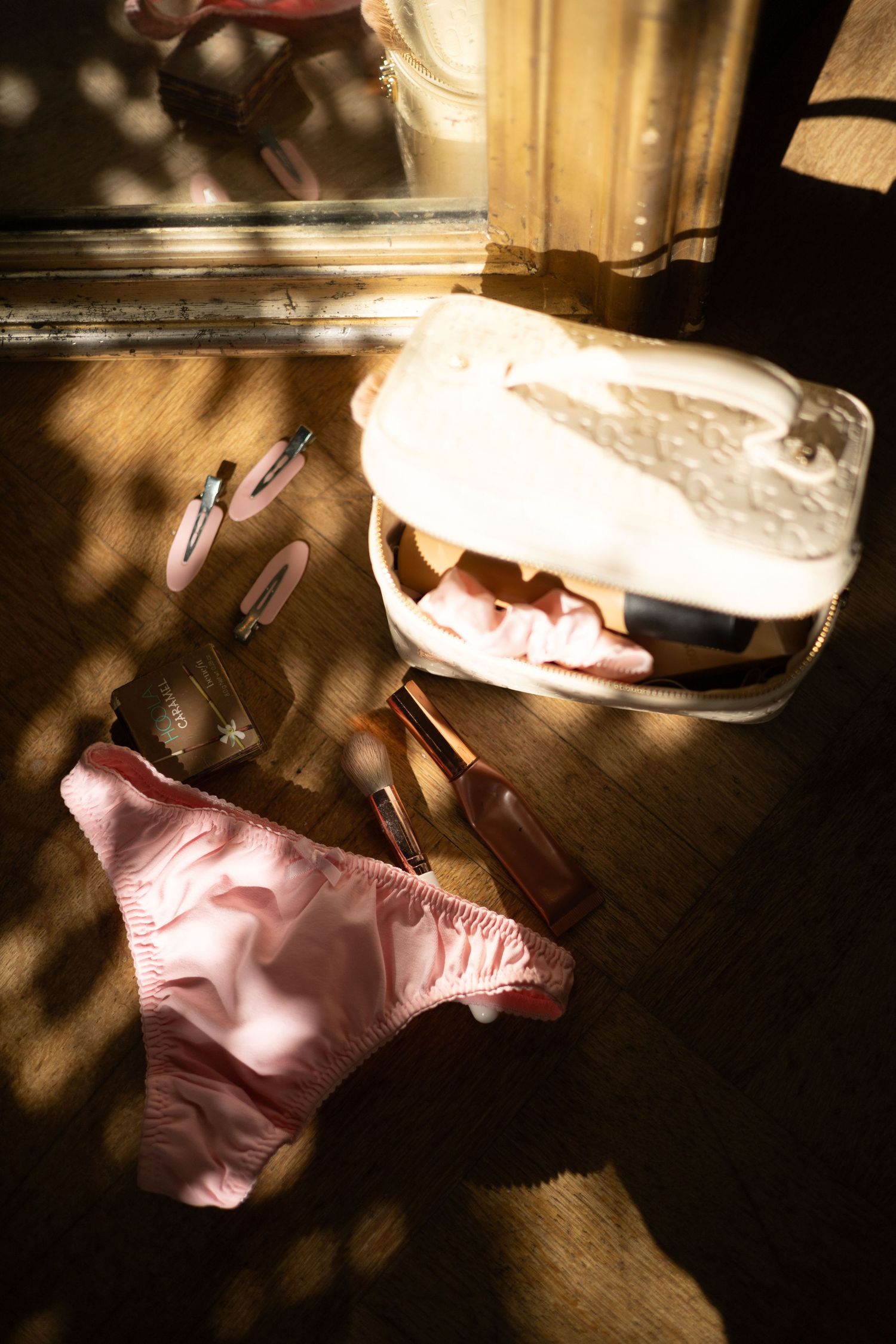Pink lingerie bottoms and makeup brushes scattered on wooden floor in sunlight.