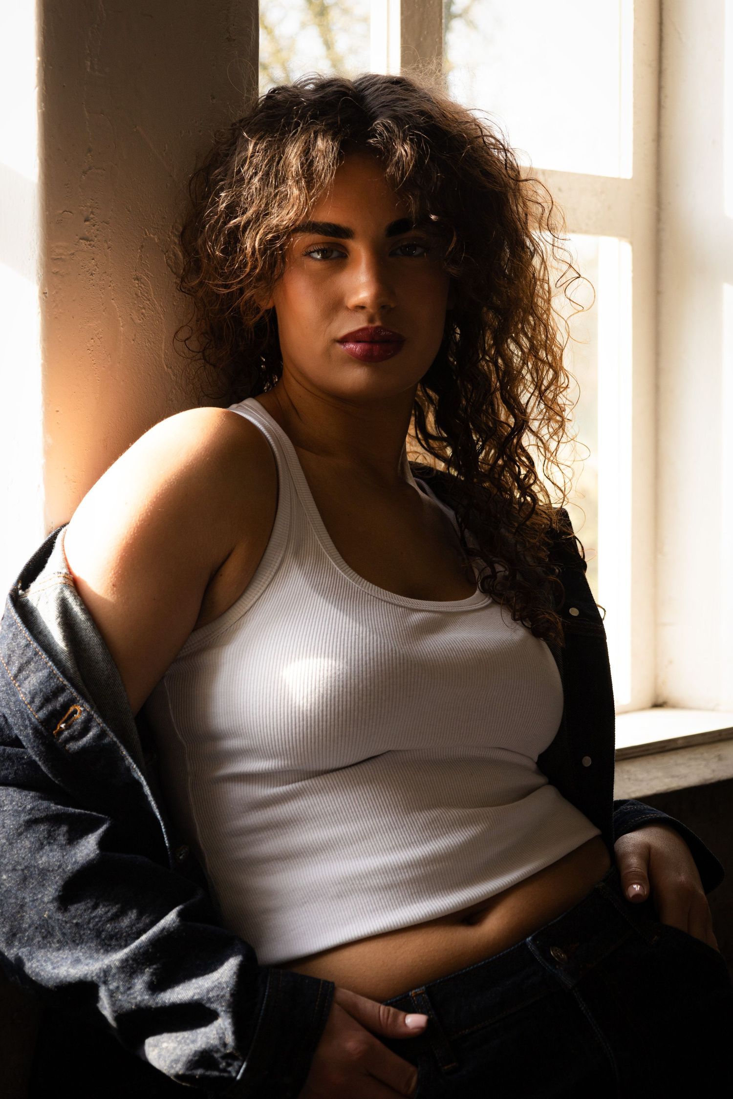 Woman with curly hair in a white tank top and denim jacket poses by a sunlit window.