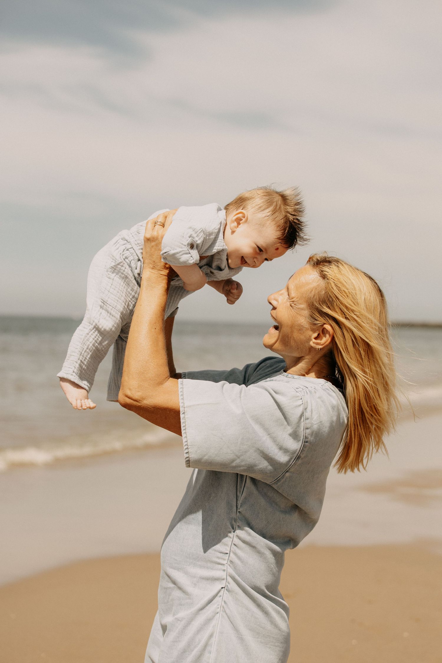 A joyful woman lifting a laughing baby high in the air on a sunny beach with ocean waves in the background.