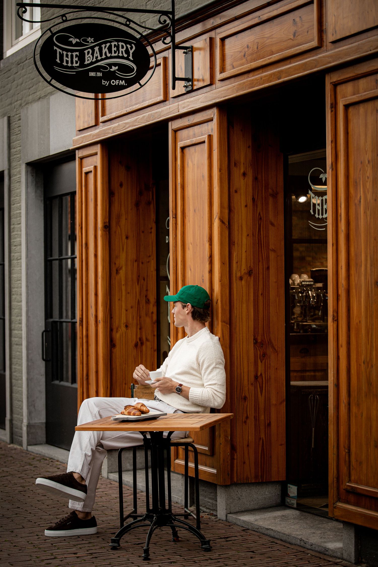 Man in white outfit and green cap enjoys breakfast outside The Bakery cafe on cobblestone street.