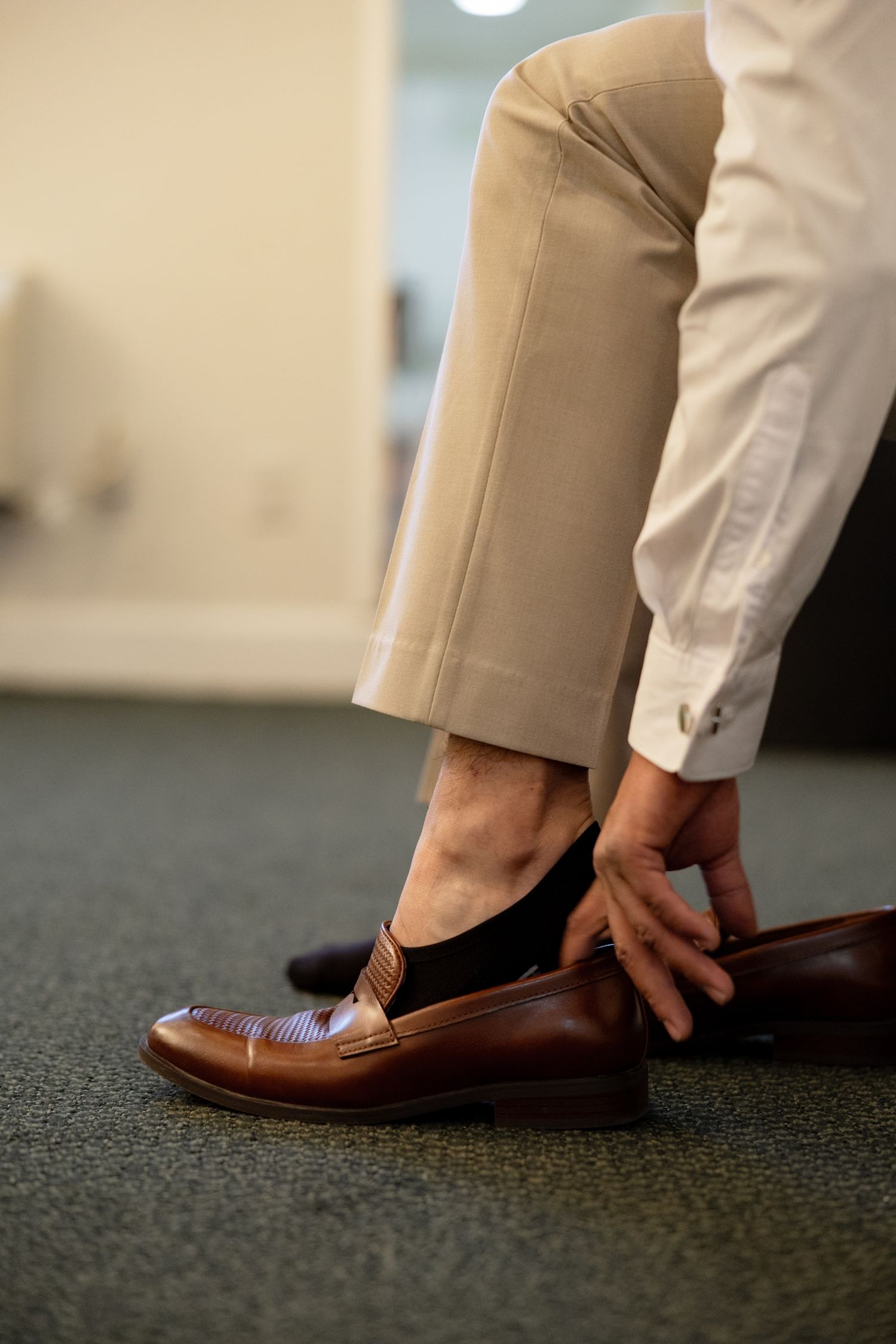 Man putting on brown leather loafer dress shoe while wearing khaki pants and white dress shirt.
