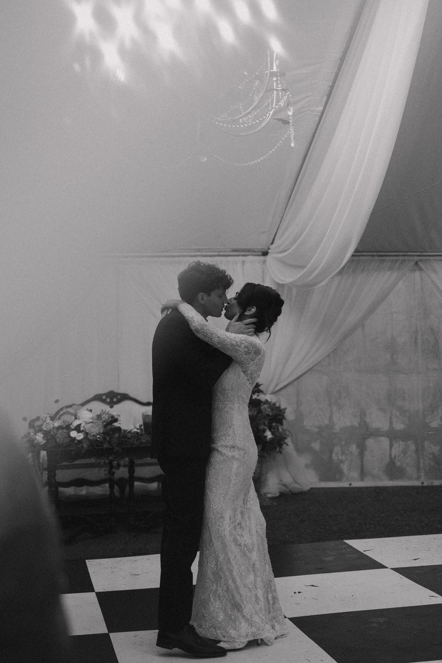 Black and white photo of bride and groom sharing first dance under chandelier on checkered floor.