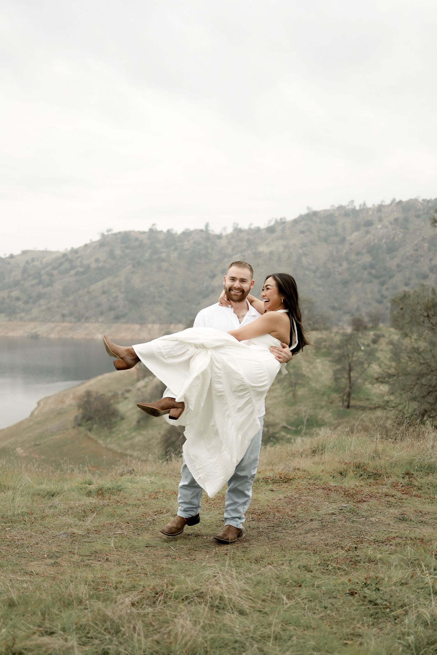 A romantic couple in white attire share a playful moment on a hillside overlooking a lake.