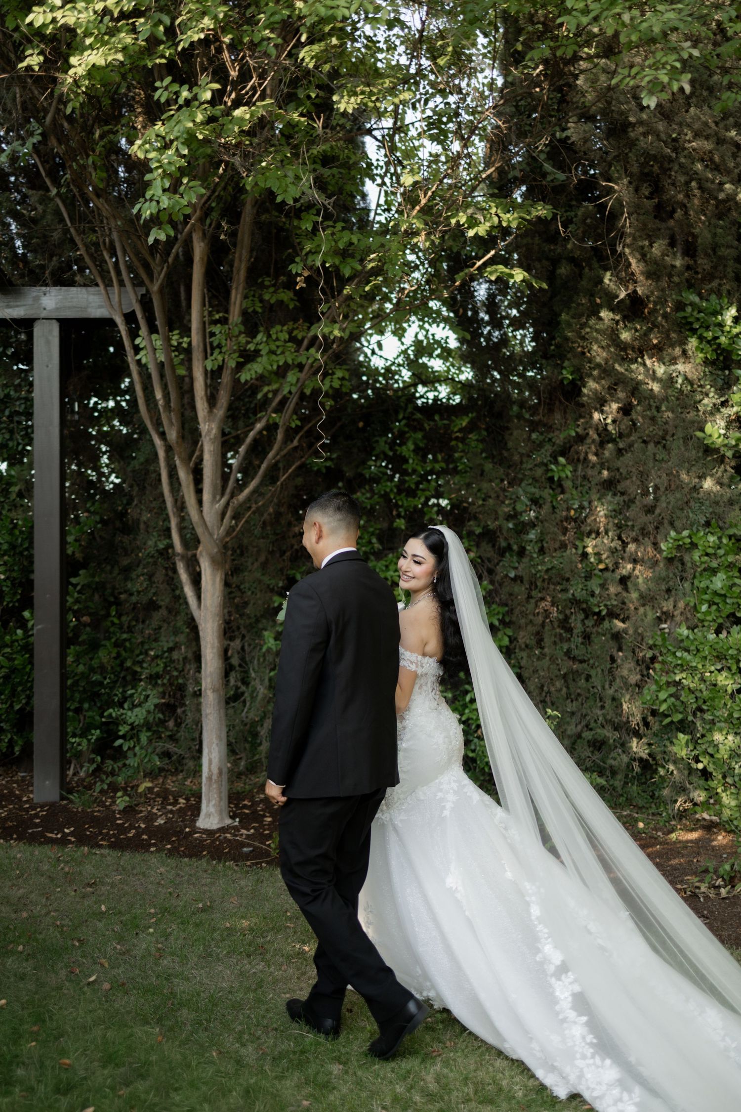 Wedding couple walking through a lush garden path with flowing white dress trailing behind.
