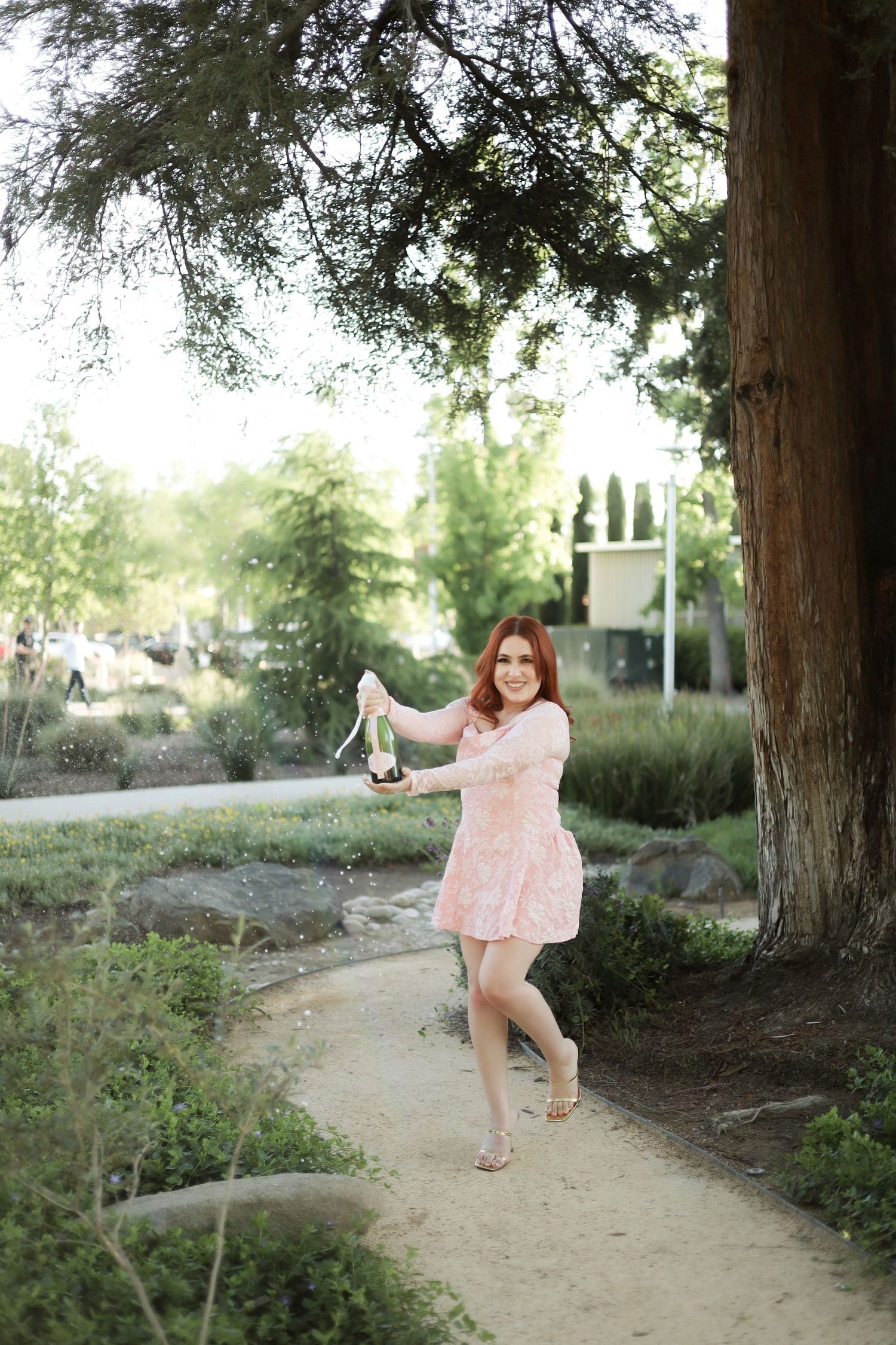 A person in a pink dress dances joyfully along a dirt path under tall trees in a natural outdoor setting.
