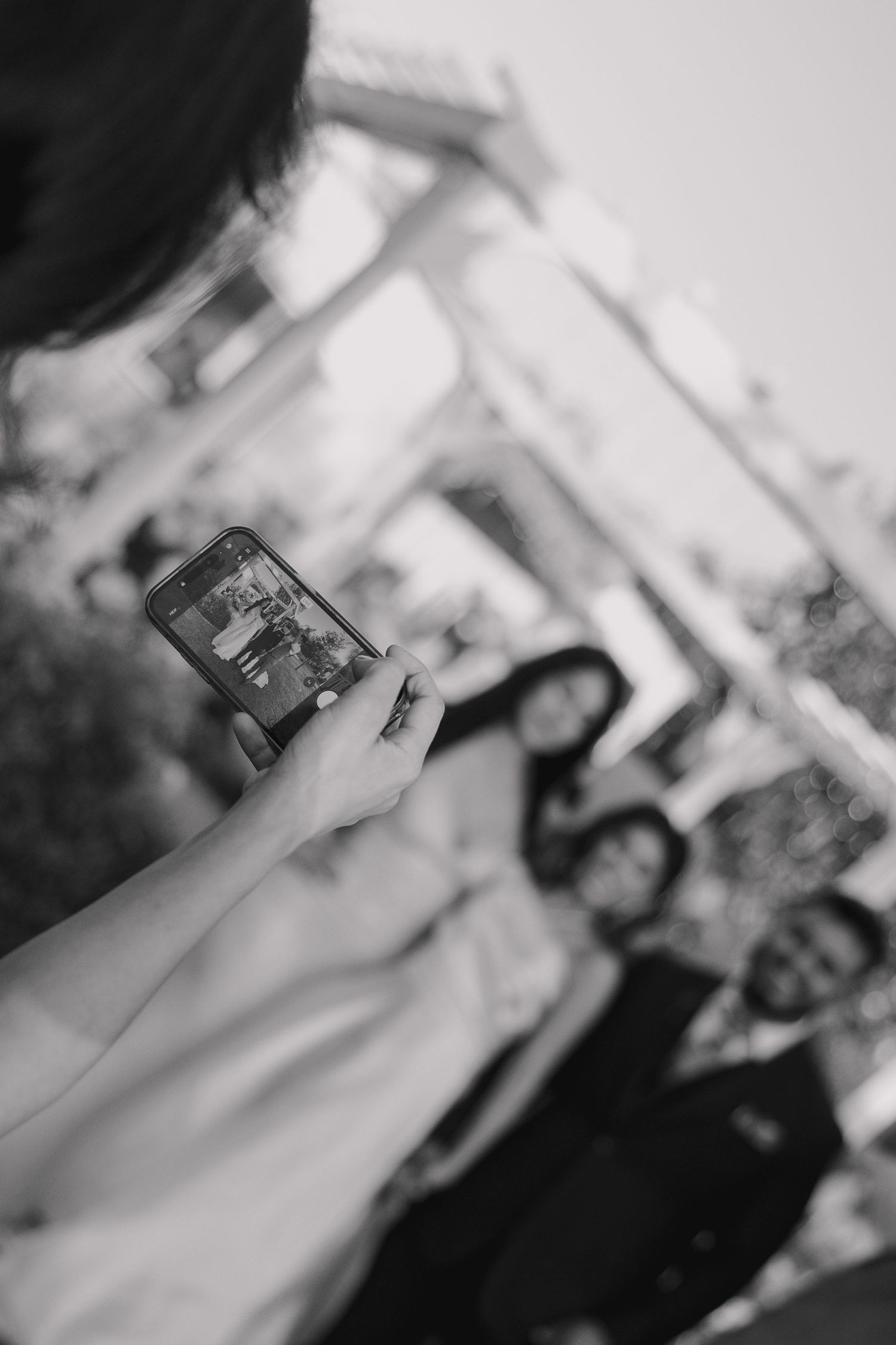 Person photographing a group of smiling wedding guests with a smartphone at an outdoor venue.