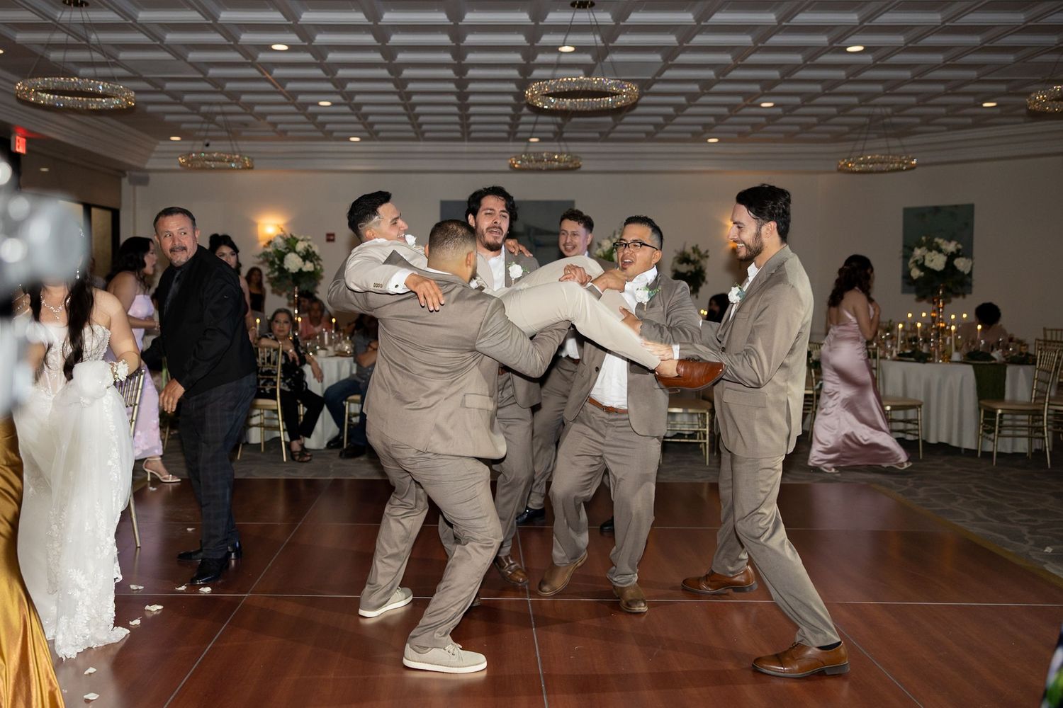 Groomsmen in gray suits lifting groom on dance floor at elegant wedding reception venue.