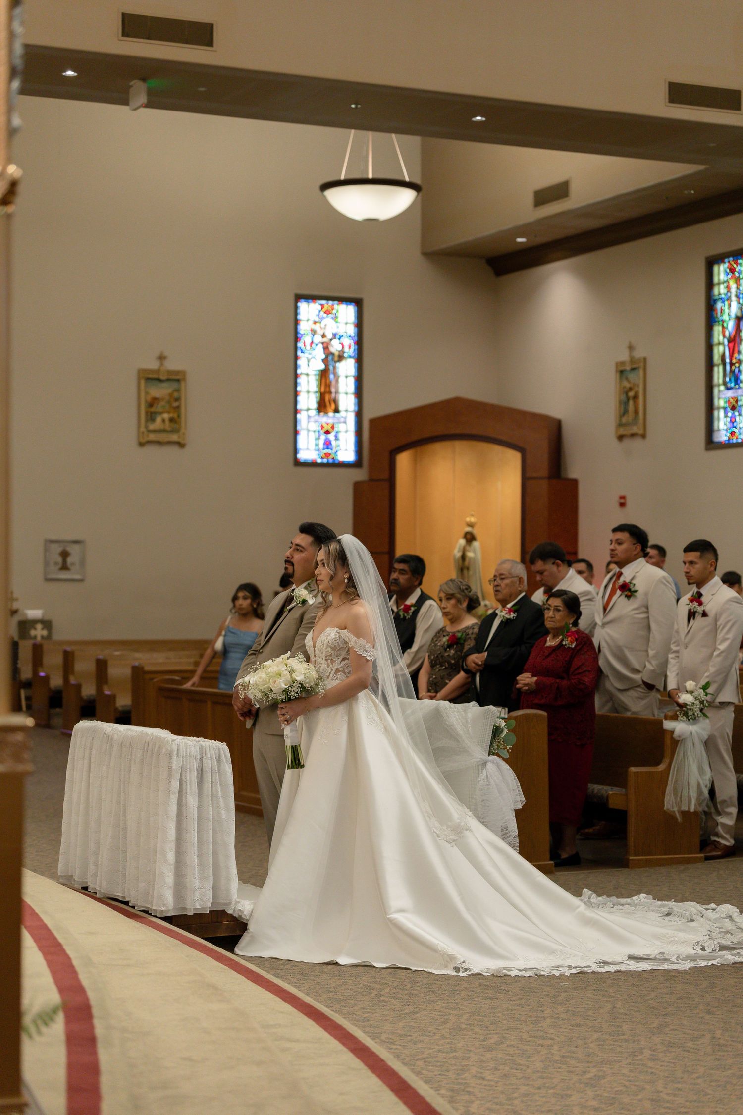 Bride in white gown with long veil stands at church altar during wedding ceremony with stained glass windows.