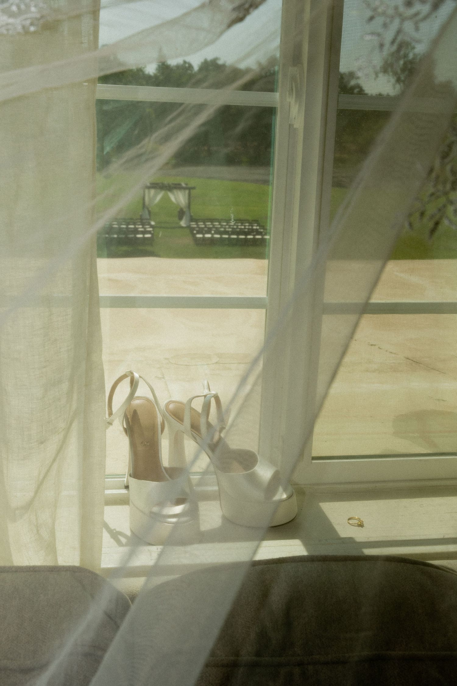 Bridal heels on windowsill with sheer curtains and outdoor wedding ceremony setup visible in background.