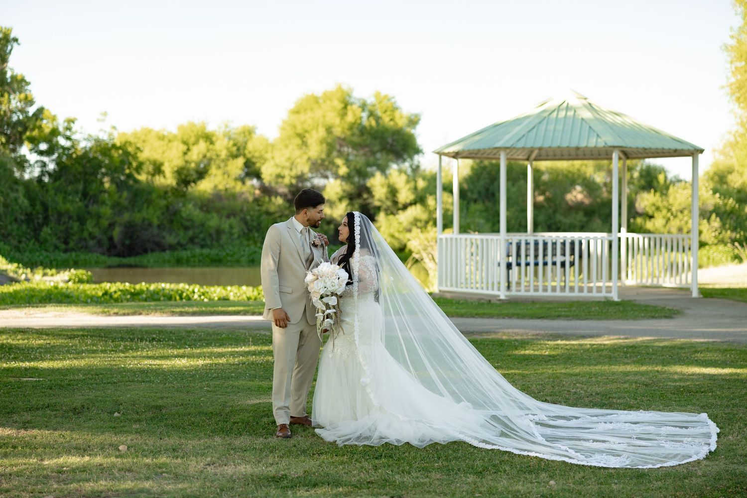 Bride and groom kissing in a lush green park with a white gazebo backdrop on their wedding day.