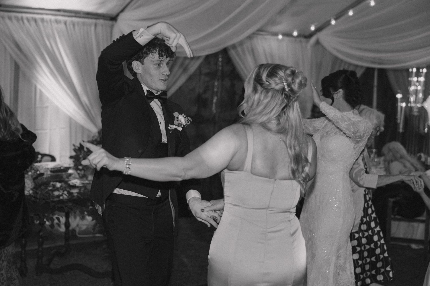 Black and white photo of groom in tuxedo dancing with guests at lively wedding reception.