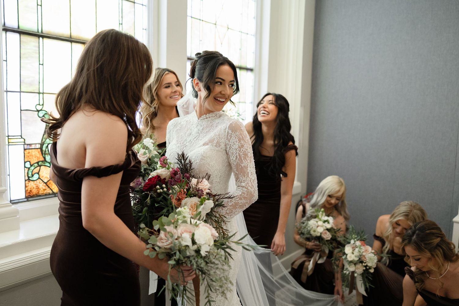 Bride in lace wedding gown surrounded by bridesmaids in dark dresses holding bouquets near stained glass window.