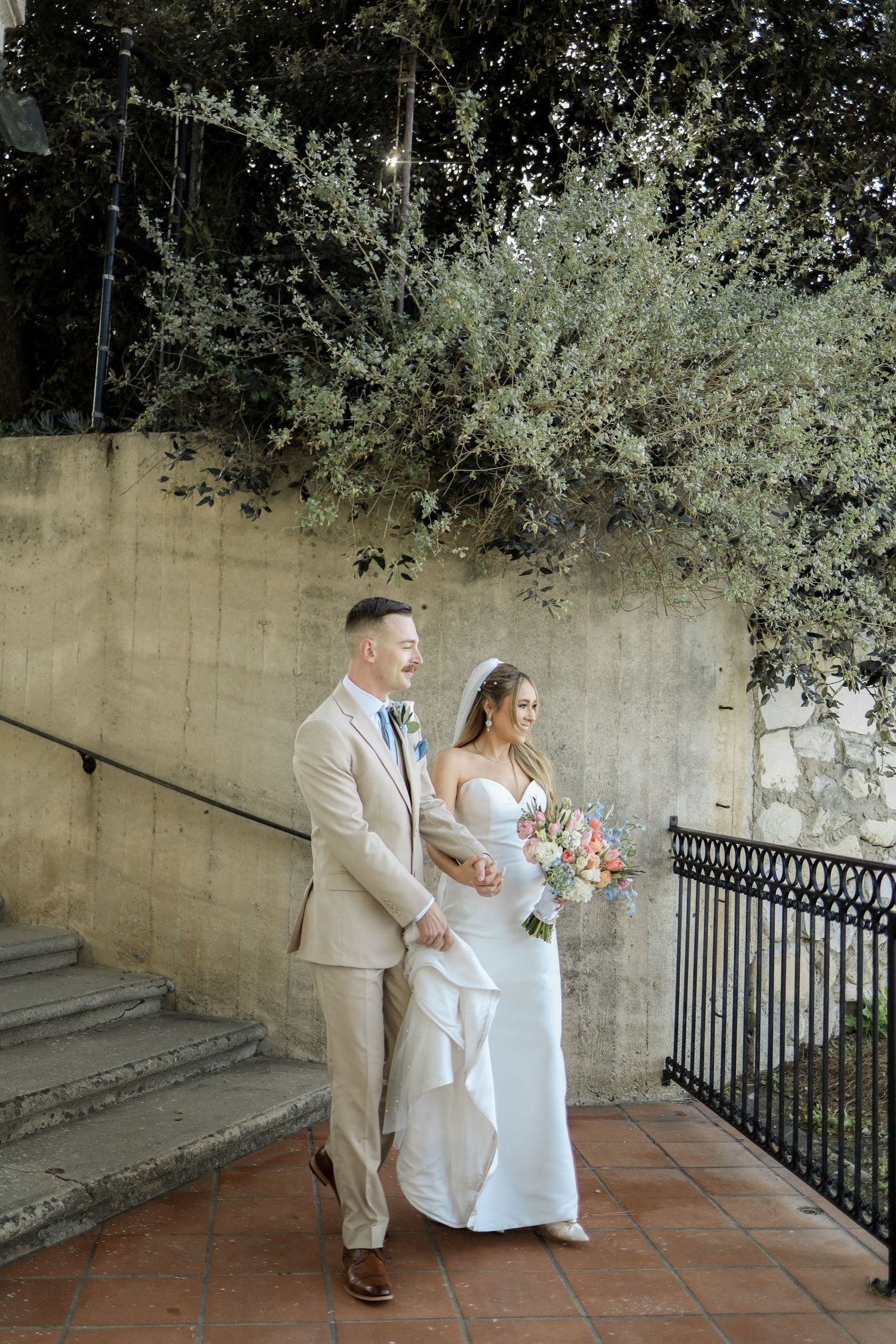 A couple in wedding attire poses together on a stone walkway with trees in the background.